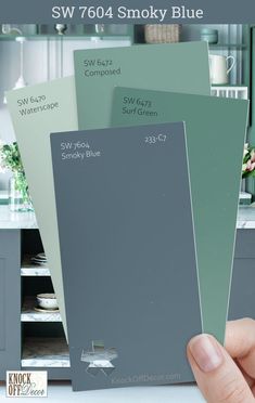 a hand holding three different shades of paint in front of a kitchen cabinet with the words sw 704 smoky blue on it