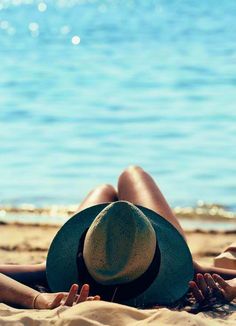 a woman laying on top of a sandy beach next to the ocean wearing a hat