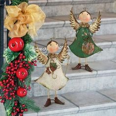 two angel figurines are standing on the steps next to a christmas wreath with holly berries and bells