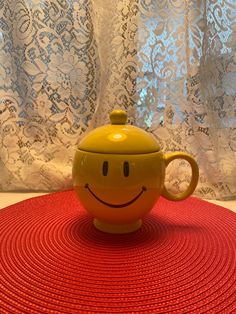 a yellow teapot sitting on top of a red mat next to a white curtain