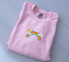 a pink t - shirt with a rainbow and clouds embroidered on the front, sitting on a white surface