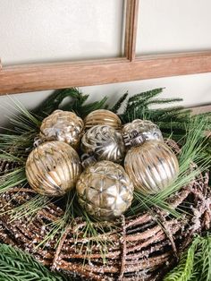 some ornaments are sitting in a basket on top of pine needles and evergreen branches, with a mirror behind them