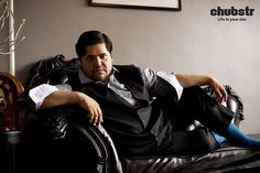 a man sitting on top of a black chair next to a wall with a clock