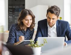 a man and woman sitting at a table looking at something on a laptop computer while another person writes in front of them