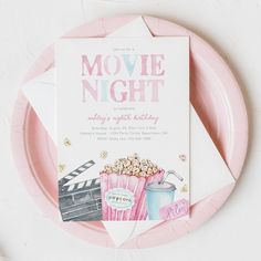 a pink paper plate with a movie night party card on it next to a cup of popcorn