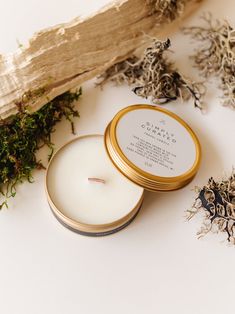 an open candle sitting next to some dried plants on a white surface with gold rimmed tin