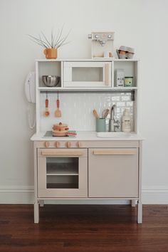 a white play kitchen with wooden floors and shelves on the wall next to an oven