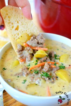 a hand holding a piece of bread over a bowl of soup with potatoes, carrots and meat