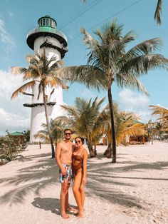 two people standing in front of a light house on the beach with palm trees behind them