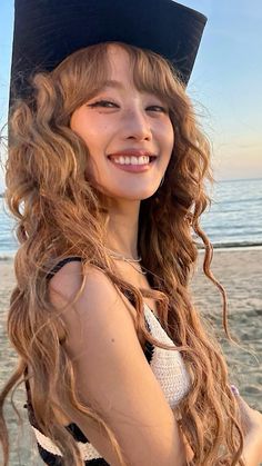 a girl with long curly hair wearing a black hat and smiling at the camera while standing in front of the ocean