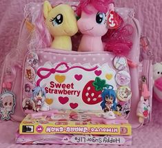 two little ponys sitting in a pink bag with other toys around them on a bed