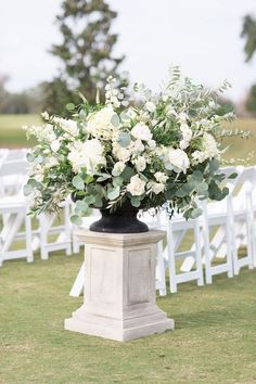 an arrangement of white flowers and greenery in a black vase on the grass at a wedding ceremony