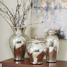 three silver vases sitting on top of a wooden table next to a painting and flowers