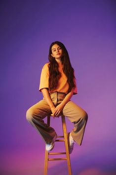 a woman sitting on top of a wooden stool in front of a purple background with her legs crossed