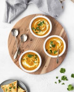 three bowls of carrot soup on a wooden plate with two slices of bread next to it