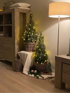 two christmas trees are sitting in baskets on the floor next to a table and lamp