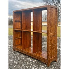 a wooden bookcase sitting on top of gravel next to a field with trees in the background