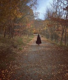 a person in a black cloak walking down a dirt road surrounded by trees and leaves