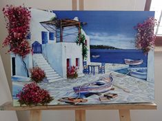 an easel with a painting of boats and flowers in front of a house by the water