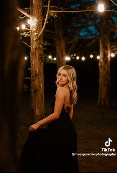 a woman in a black dress standing next to some trees at night with lights strung overhead