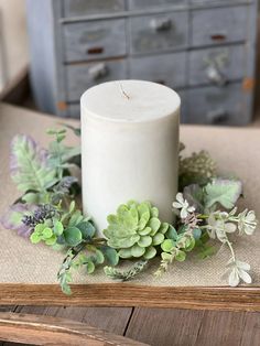 a white candle sitting on top of a wooden table next to flowers and greenery