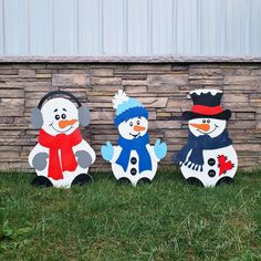 three snowmen are standing in the grass near a brick wall and stone wall behind them