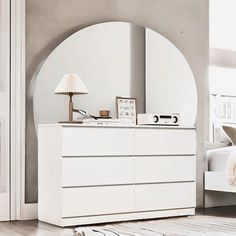 a white dresser sitting next to a bed in a bedroom under a large round mirror