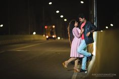 a man and woman standing next to each other on a bridge at night with street lights in the background