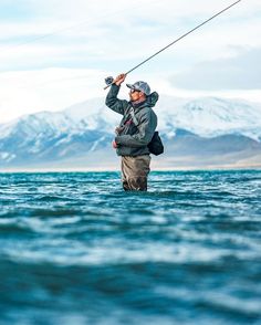 a man standing in the water while holding onto a fishing line with mountains in the background