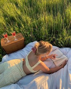 a woman laying on a blanket reading a book and drinking wine in front of some tall grass