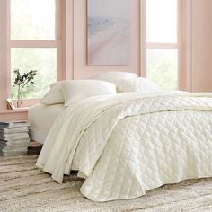 a bed with white comforter and pillows in a room next to two large windows