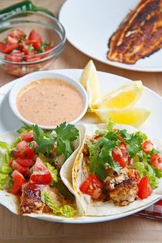 two tacos with lettuce, tomatoes and sauce on a plate next to lemon wedges