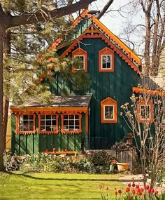 a green house in the woods with flowers and trees around it's front yard