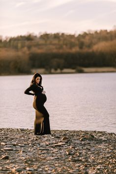 a pregnant woman standing on the shore of a lake wearing a long black and tan dress