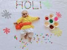 a baby laying on the ground next to paint bowls and paper flowers that spell out holi