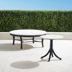 two black and white tables sitting on top of a cement floor next to a hedge