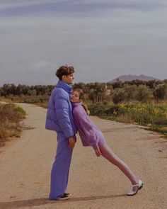 a man and woman standing on the side of a dirt road in front of mountains