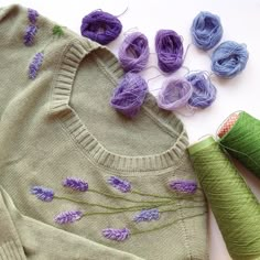 some knitting supplies are laying out on the floor next to yarn and needles, including one green sweater with purple flowers