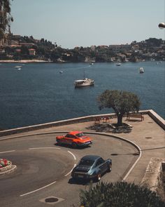 two cars are driving on the road by the water with boats in the bay behind them