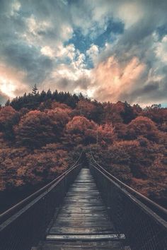 a wooden bridge with trees in the background and cloudy skies above it on a sunny day