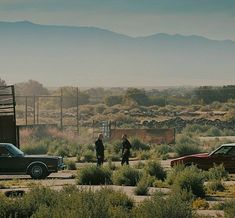 two men are standing in the middle of an open field with cars parked on it