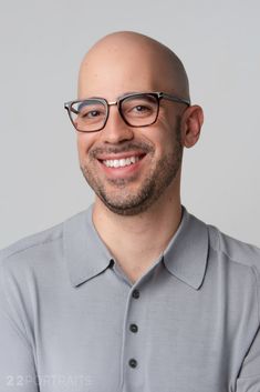 a bald man wearing glasses and smiling for the camera in front of a gray background