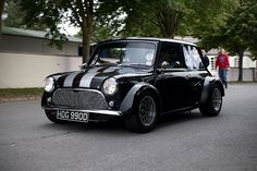 an old black mini car driving down the street with people standing around it and trees in the background