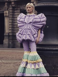 a woman standing on top of a street next to a black trash can wearing a purple and green dress