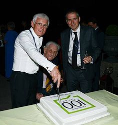 two men are cutting into a large white cake with the number 60 written on it