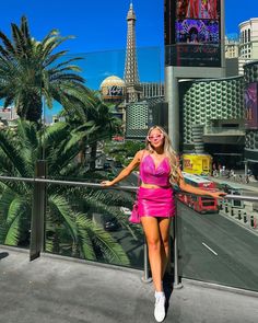 a woman in a pink dress standing on a balcony with palm trees and buildings behind her