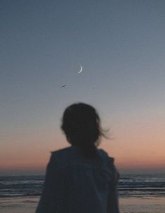 a person standing in front of the ocean watching the moon rise over the water at sunset