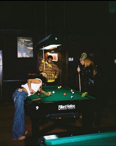 some people are playing pool in a dark room with lights on and one person is leaning over the pool table