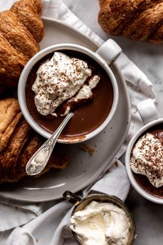 two cups of hot chocolate with whipped cream and croissants on the side