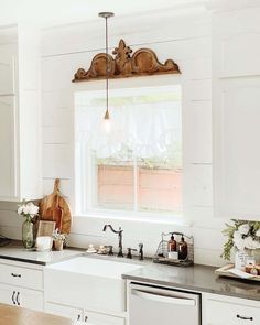 Admire this farmhouse kitchen surrounded by white shiplap walls with a sink by the window. Mounted above the window is a beautiful carved wooden piece that adds a rustic touch to the view, which chose by the stacked wooden cutting board on the counter below. Decorate the countertop with white flowers to add a natural, earthy vibe...   Image: whitecottagecreek